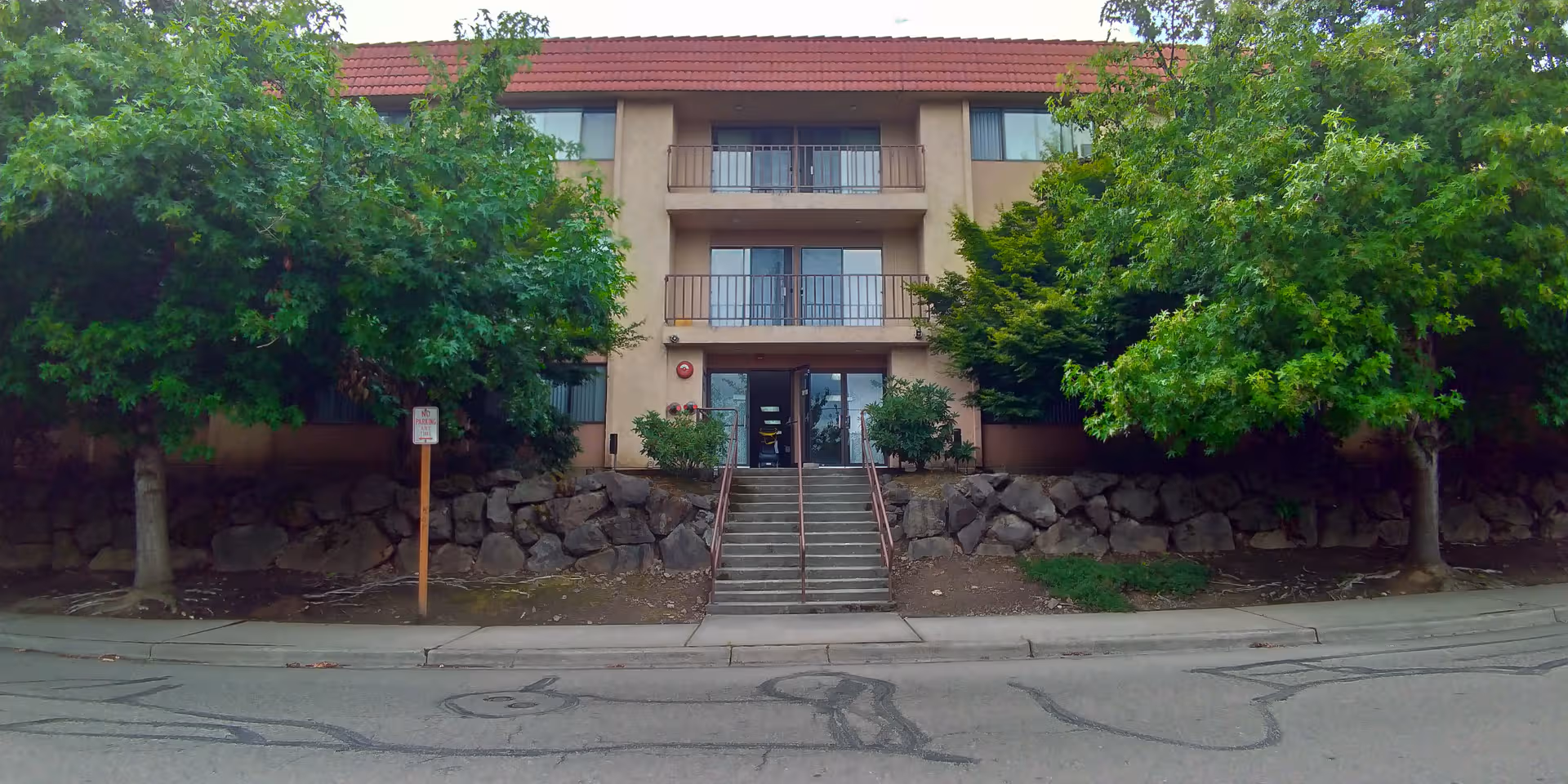 Exterior view of a beige, multi-story senior living building framed by trees and a sidewalk.