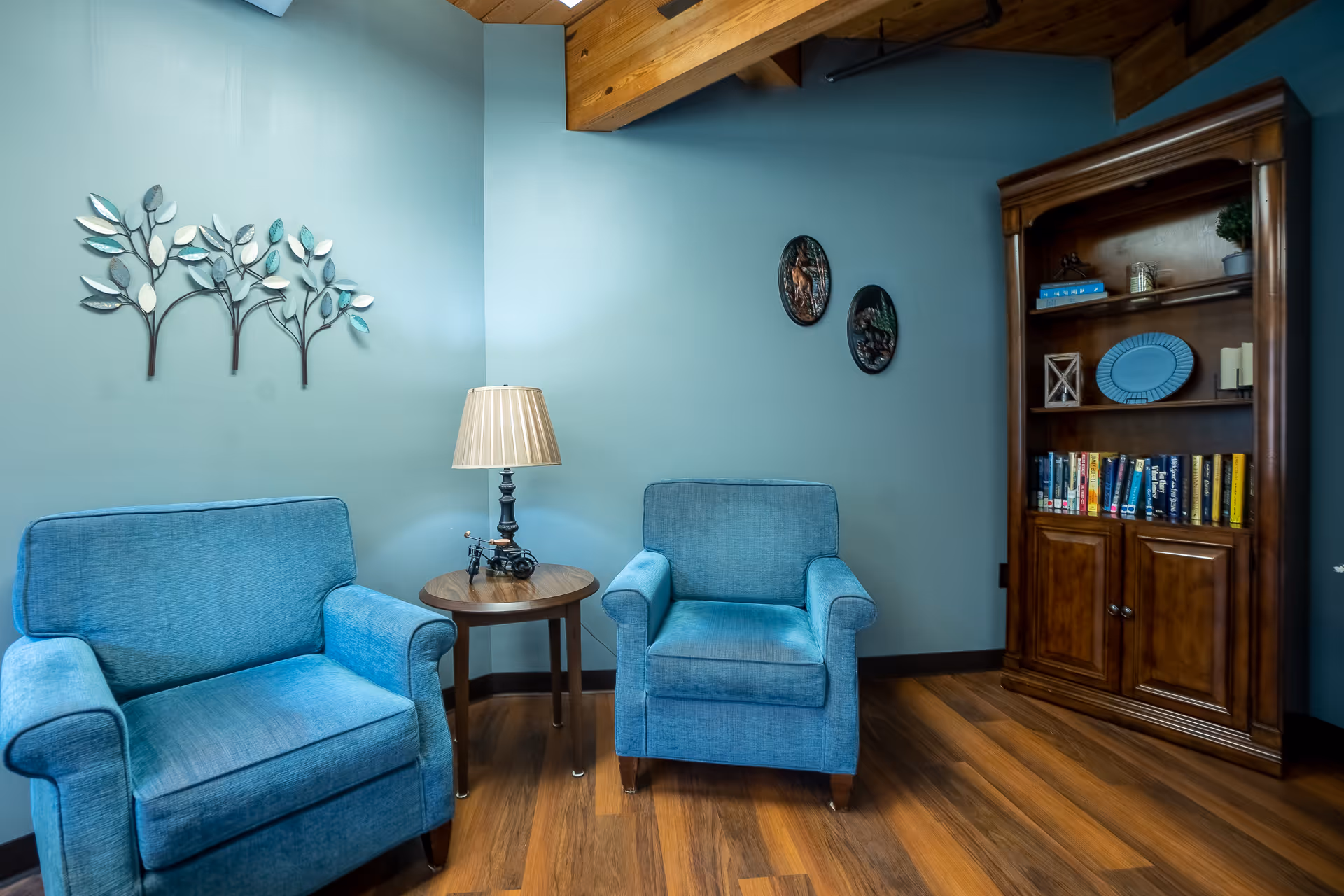 A cozy sitting area with two blue upholstered armchairs separated by a round wooden side table with a lamp. The walls are painted light blue and decorated with metal leaf wall art and two oval plaques. A wooden bookshelf filled with books and decorative items stands against the wall on the right. The floor is wood and the ceiling has exposed wooden beams.