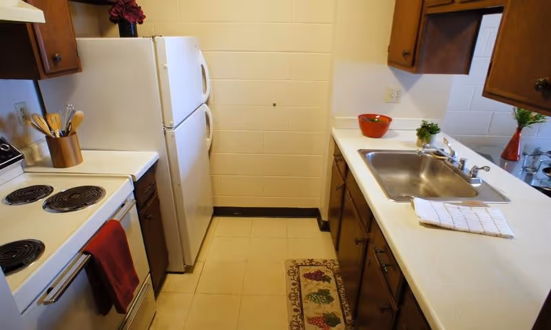A compact kitchen with white countertops and wooden cabinets. The kitchen features a white electric stove with a red towel hanging on the handle, a white refrigerator, a stainless steel sink with a white dish towel, and small decorative plants and bowls on the counters. The floor has beige tiles and a small rug with a grapevine pattern.