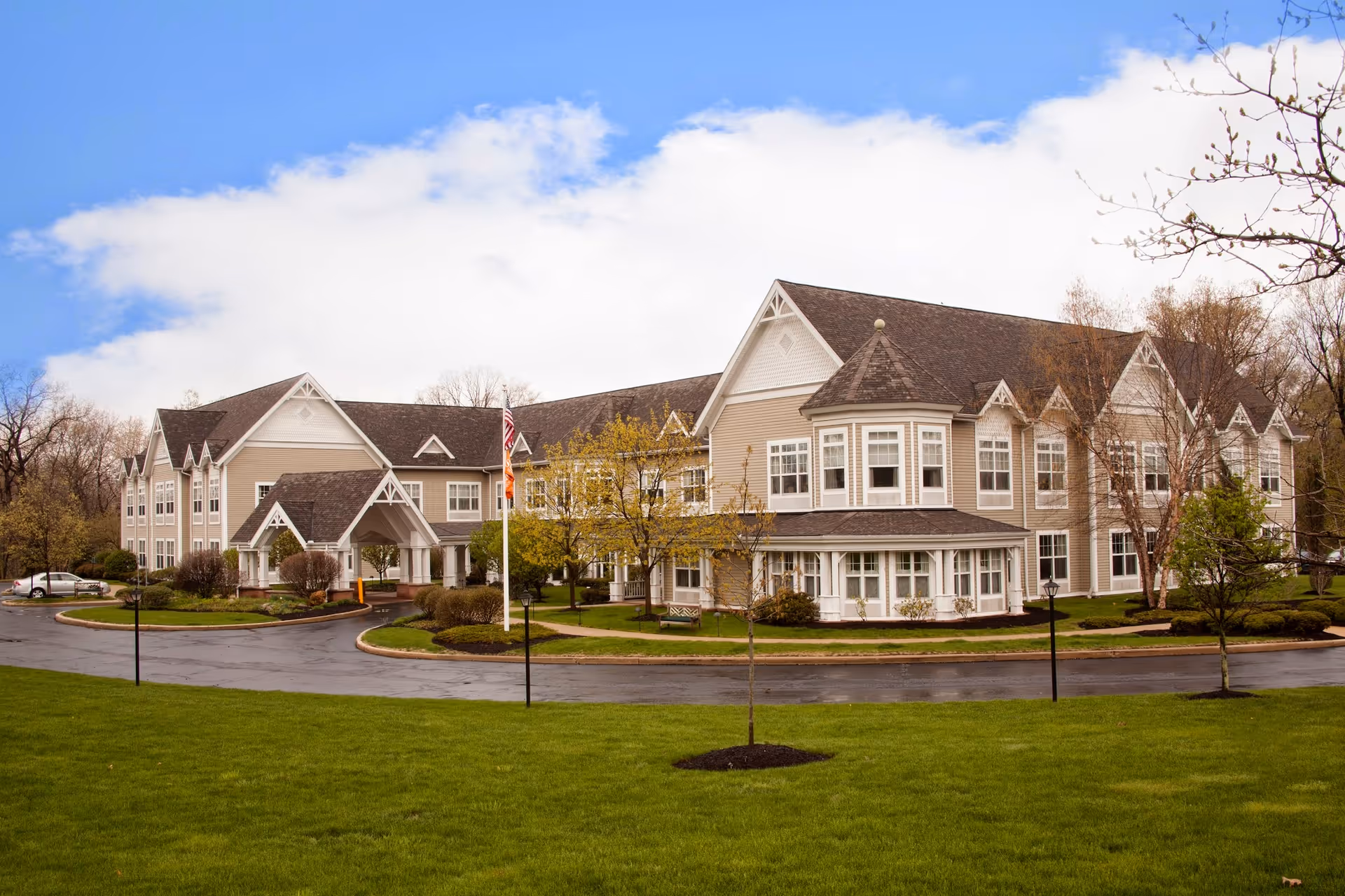 Exterior view of a large, two-story senior living facility building with beige siding and multiple gabled roofs. The building is surrounded by well-maintained green lawns, small trees, and shrubs. A paved driveway curves in front of the entrance, and an American flag is visible near the entrance. The sky is partly cloudy with patches of blue.