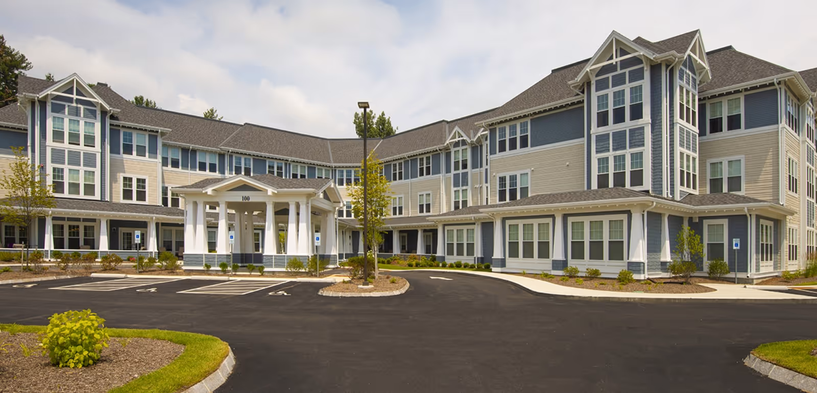 Front exterior of a three-story senior living residence with a covered entrance, parking lot, and landscaped grounds.