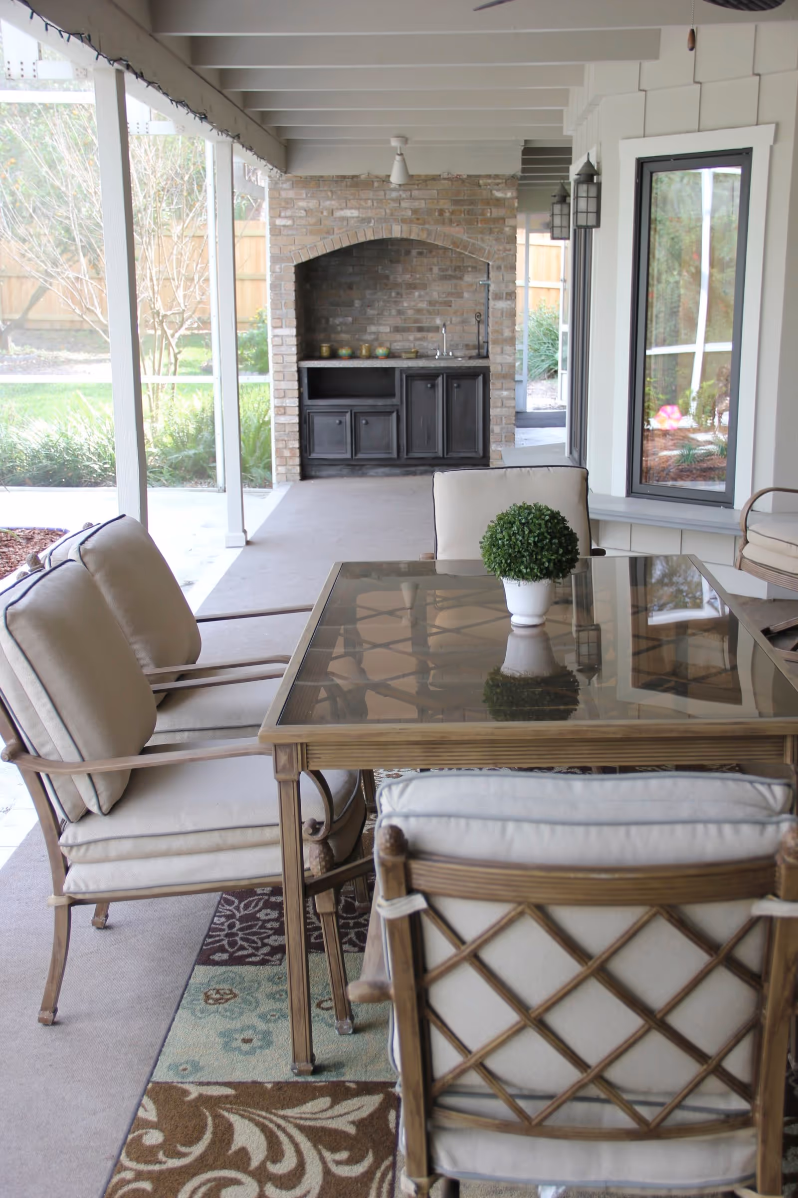 Covered outdoor patio area with a glass-top table surrounded by cushioned chairs. A small potted plant is centered on the table. In the background, there is a brick built-in counter with cabinets and a small sink. The patio overlooks a garden with grass and trees, enclosed by a wooden fence.
