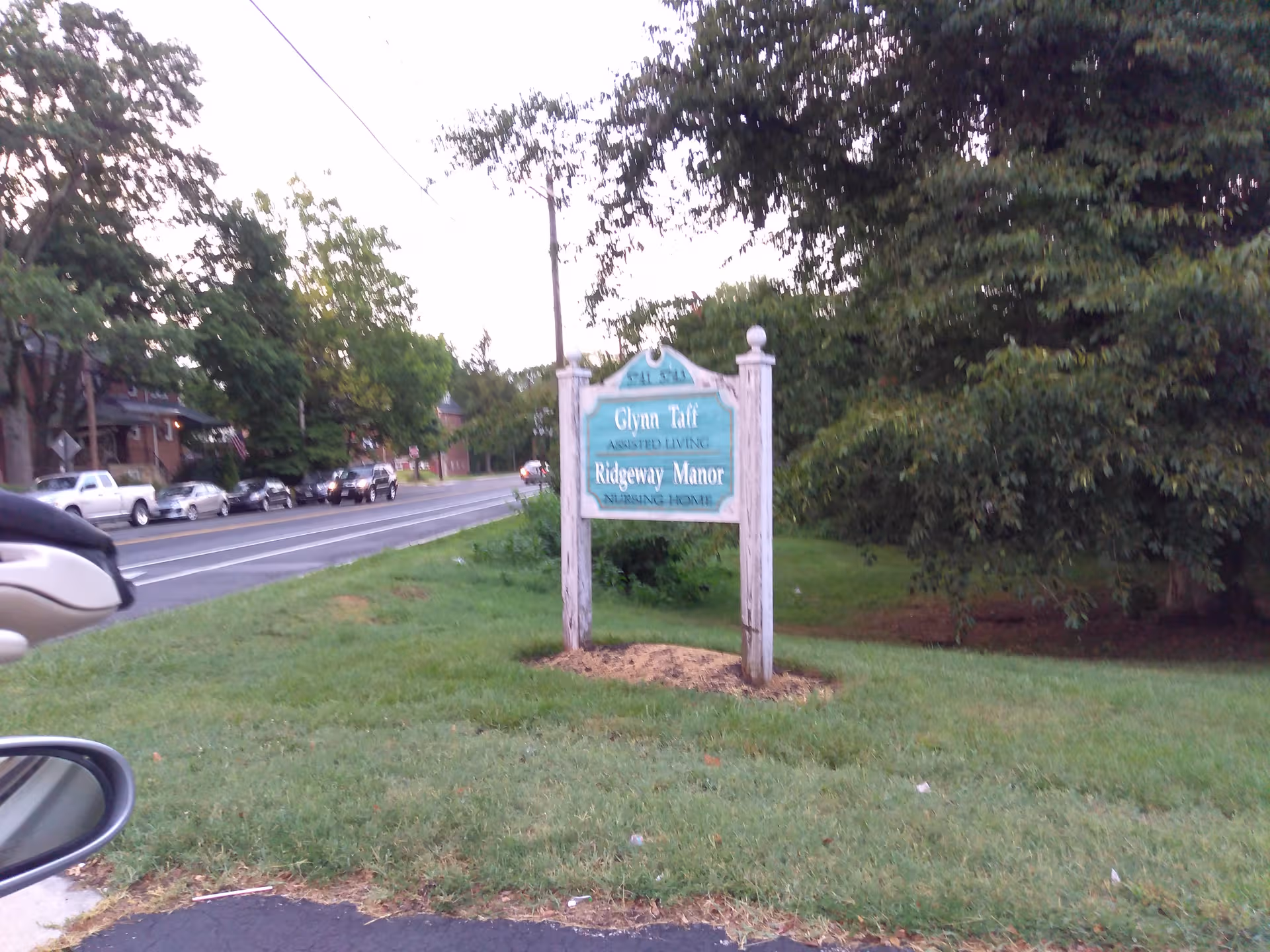 Street view with a blue Ridgeway Manor sign on a grassy verge beside a tree-lined road