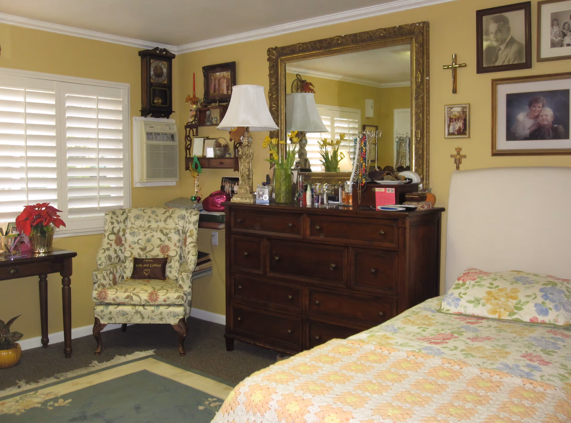 A cozy bedroom with a floral bedspread, wooden dresser topped with a lamp and large mirror, and a patterned armchair by a shuttered window.