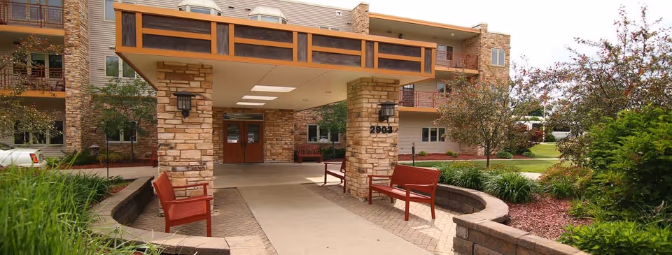 Entrance of a senior living facility with a covered drop-off area supported by stone pillars. There are red benches on either side of the walkway, surrounded by landscaped greenery and trees. The building has multiple floors with balconies and windows.