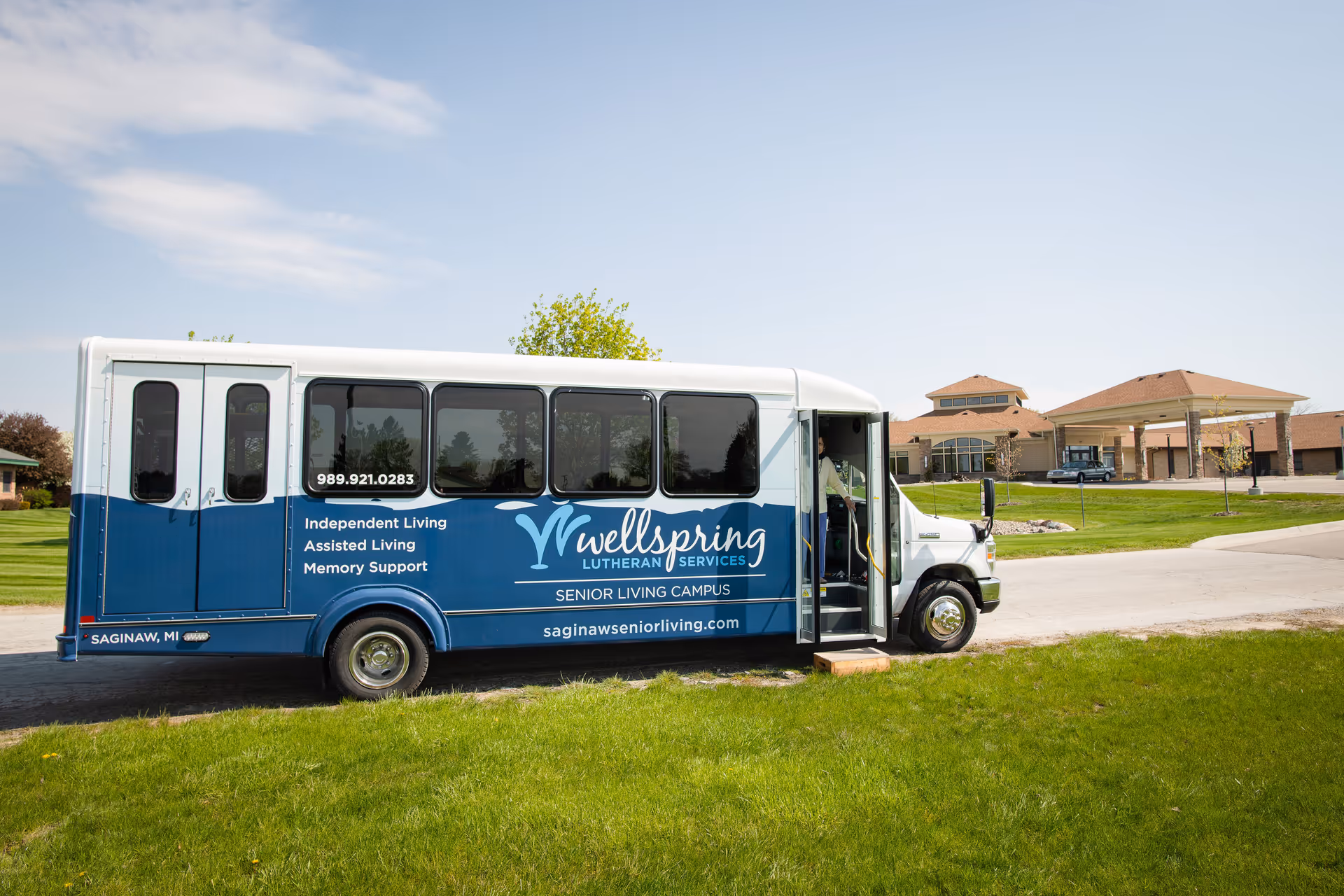 A blue and white shuttle bus parked on a driveway in front of a senior living campus building. The bus has the Wellspring Lutheran Services logo and text advertising Independent Living, Assisted Living, and Memory Support services. The building in the background has a covered entrance and is surrounded by green grass and trees under a clear sky.