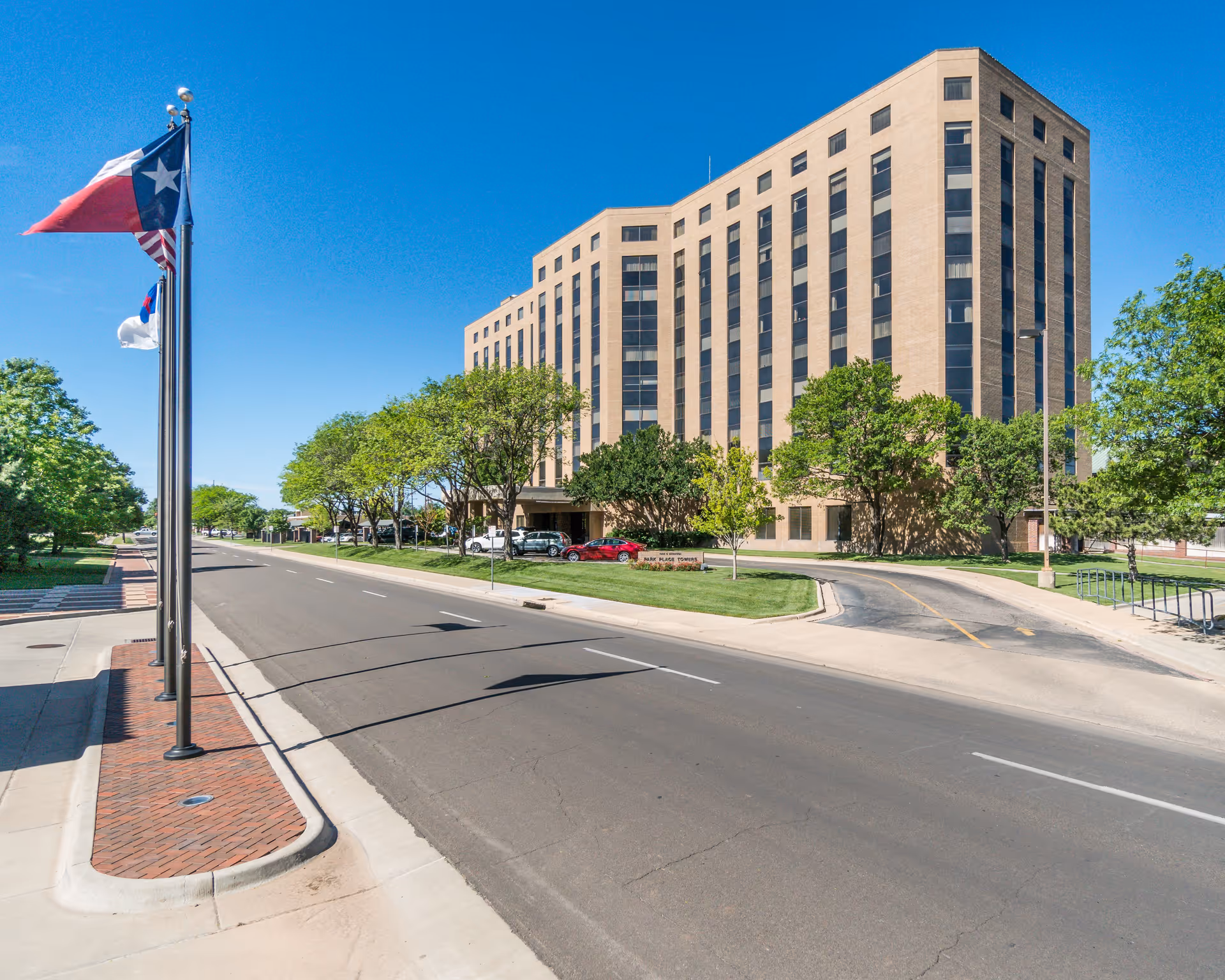 Exterior view of a multi-story beige brick building named Park Place Towers at Park Central, with a clear blue sky, several trees, and a road with flagpoles displaying the Texas state flag, the American flag, and another flag.