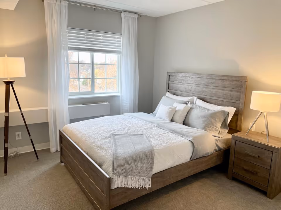 A neatly made bed with gray and white bedding in a bedroom. The bed has a wooden headboard and footboard. There is a wooden nightstand with a lamp on the right side of the bed and a tall floor lamp on the left side near a window with white curtains and blinds. The walls are painted light gray and the floor is carpeted.