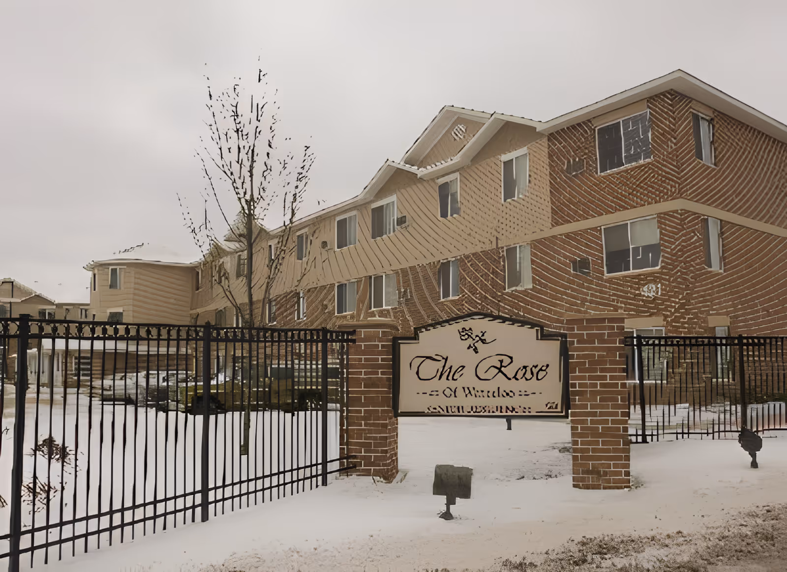 Snow-covered exterior of a multi-story residential building with a wrought-iron fence and a brick sign that reads "The Rose".