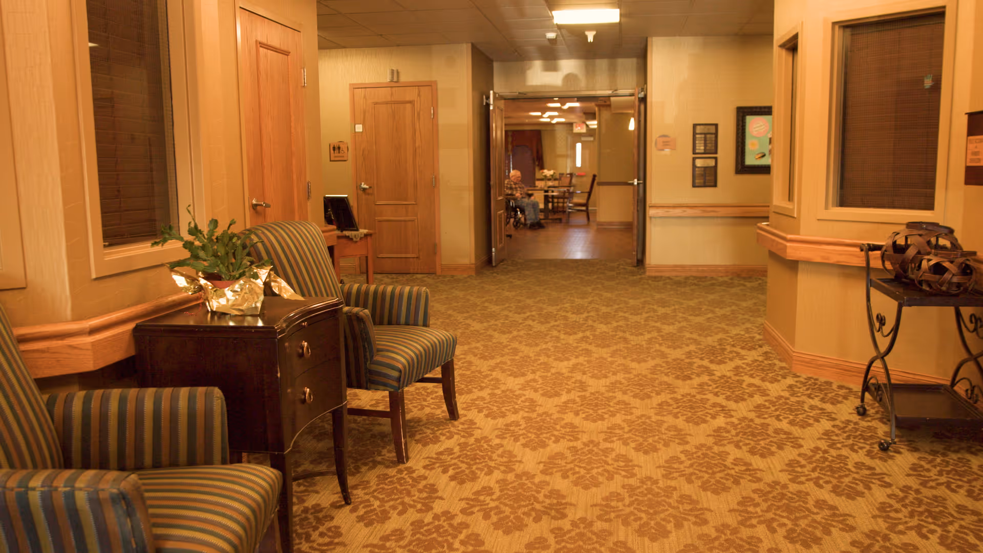 A warmly lit hallway in a senior living facility with patterned carpet and beige walls. There are two striped armchairs and a small dark wooden table with a potted plant on the left side. The hallway leads to a dining area where a person is seated at a table. On the right side, there is a small decorative metal cart with an ornamental object on top.