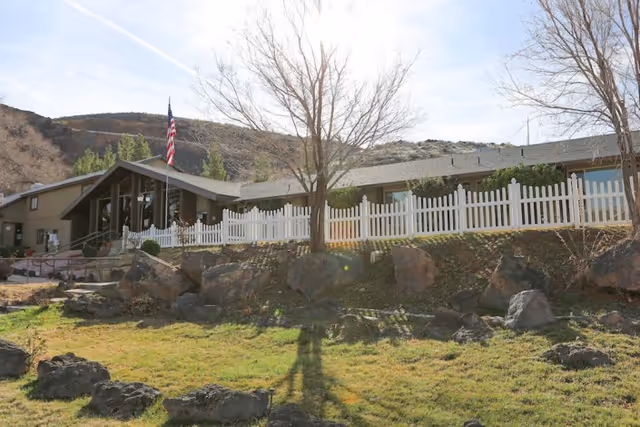 Front exterior of a one-story health and rehabilitation building with a white picket fence, rock landscaping, and an American flag.