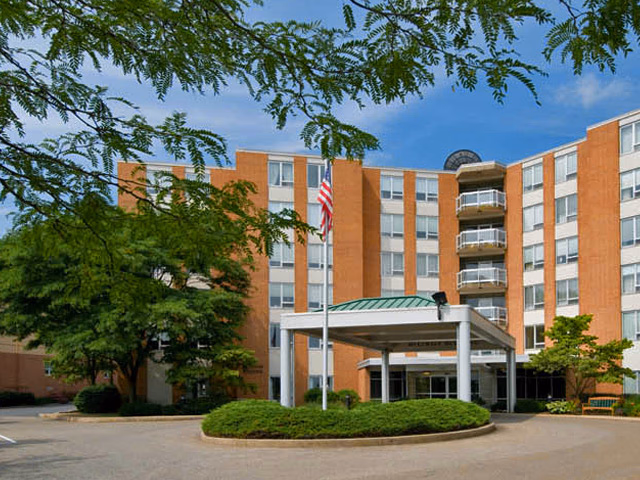 Exterior view of a multi-story brick building with balconies and large windows, surrounded by trees and greenery. There is a covered entrance with white pillars and an American flag on a flagpole in front of the building.