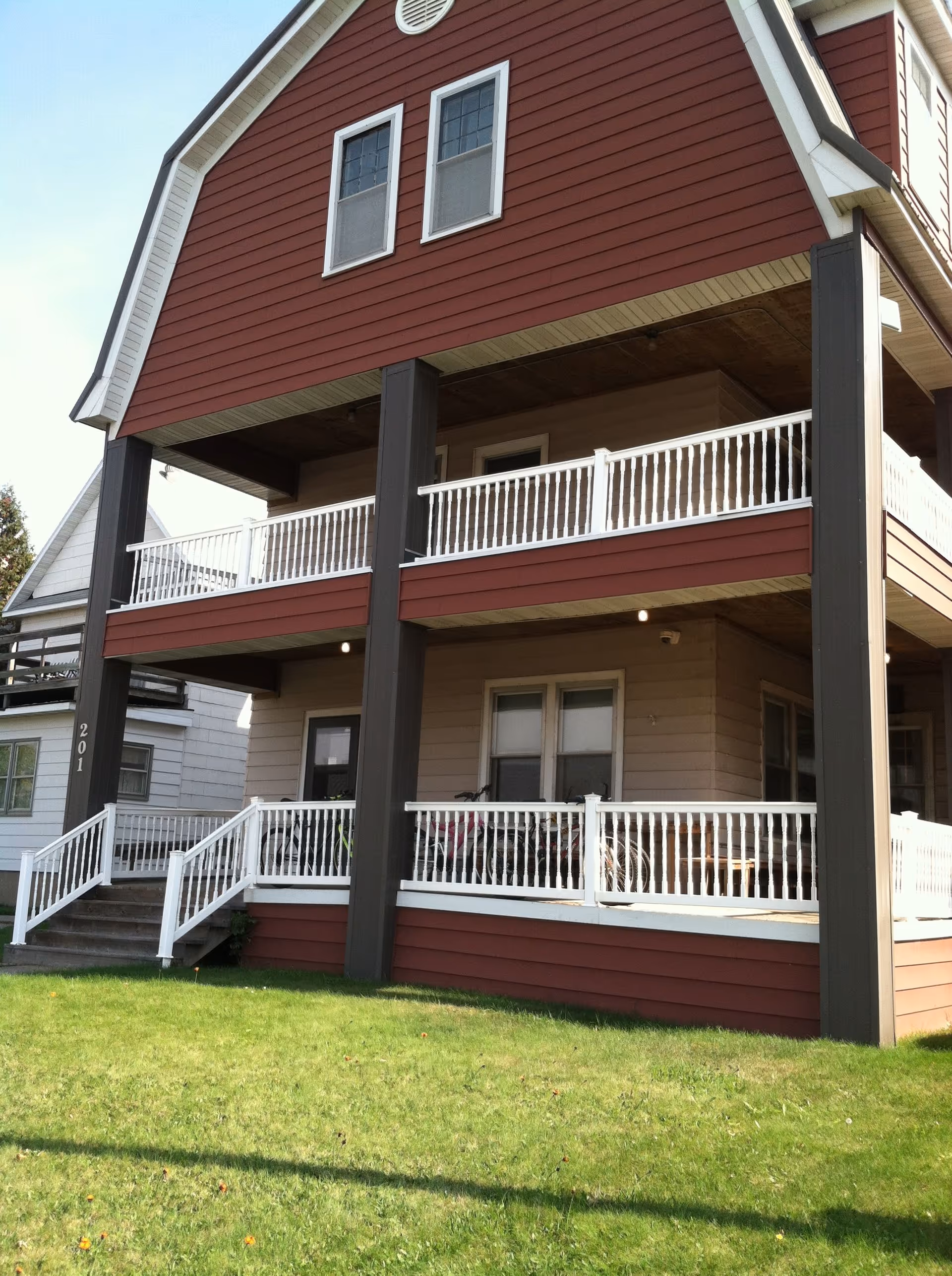 Exterior view of a two-story residential building with red siding and white railings on the balconies and porch. The building has a green lawn in front and the number 201 is visible on one of the dark brown support pillars.