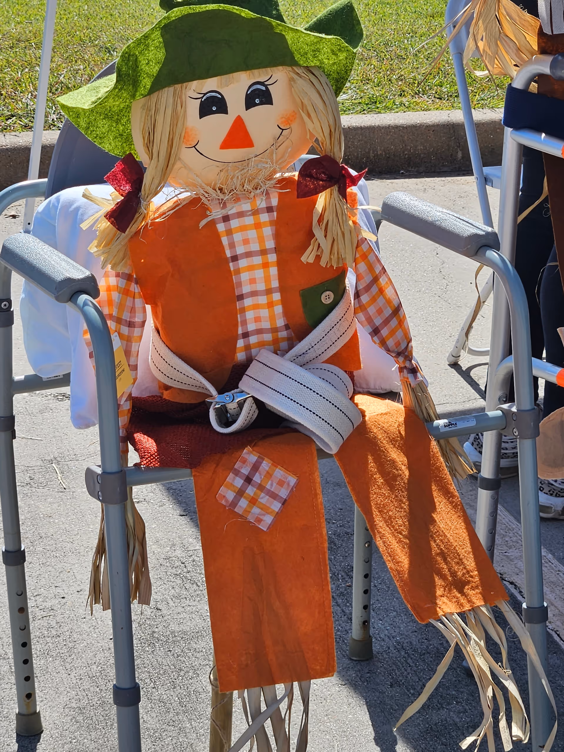 A colorful scarecrow decoration sits on a walker outdoors beside chairs.