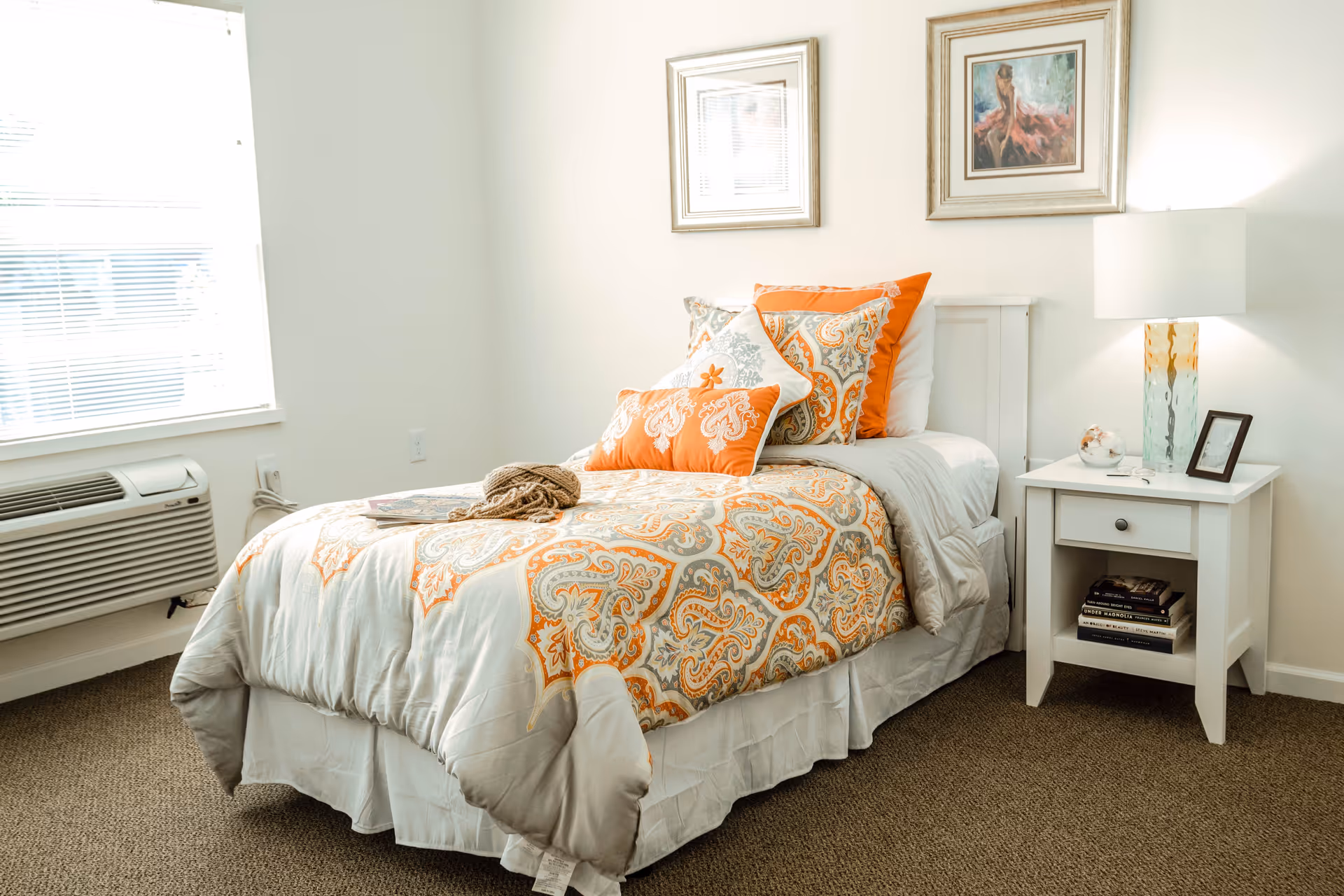 A neatly made single bed with a white, orange, and gray patterned comforter and multiple decorative pillows in orange and white. Next to the bed is a white nightstand with a drawer and an open shelf holding books. On top of the nightstand is a glass lamp with an orange and green base, a small glass bowl with seashells, and a framed photo. Two framed paintings hang on the wall above the bed. A window with blinds and an air conditioning unit are visible on the left side of the room.