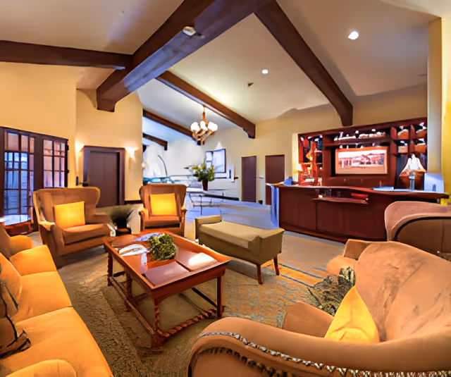 Cozy senior living lounge with sofas and armchairs around a coffee table, a vaulted beamed ceiling, and a reception/bar area in the background.