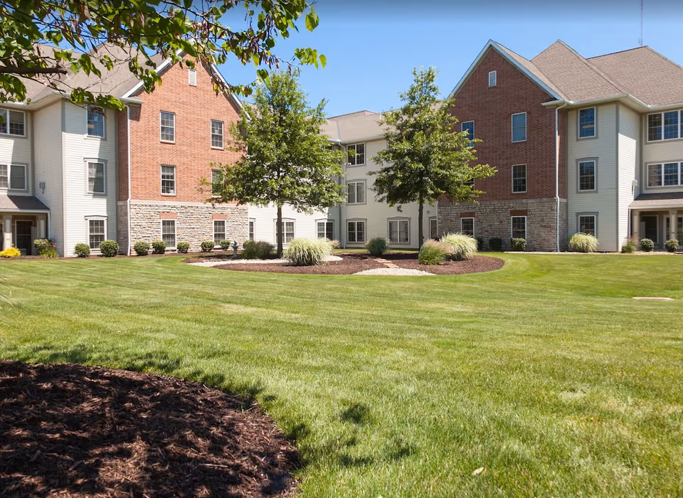 Front view of a multi-story brick-and-siding senior living building with a large manicured lawn and trees.