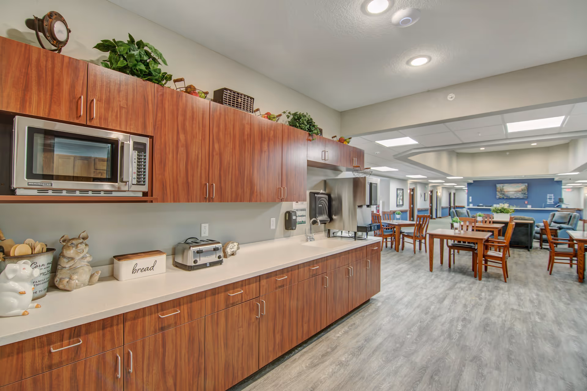 Interior view of a senior living facility kitchen and dining area featuring wooden cabinets with a microwave, toaster, and decorative items on the countertop. The space opens into a dining area with wooden tables and chairs, and a lounge area with armchairs and a blue accent wall with a painting.