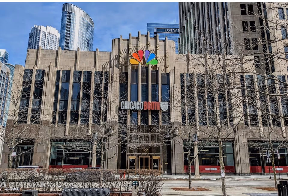 Urban office building facade with Chicago Booth signage, leafless trees, and a colorful NBC logo on top.