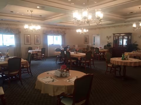 Senior living dining room with round tables, chairs, and chandeliers in soft lighting.