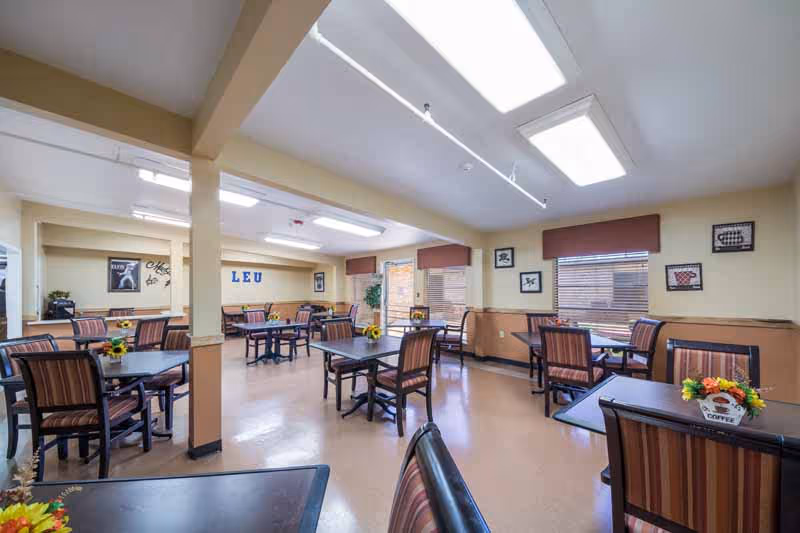 Sunlit senior living dining hall with multiple tables, striped chairs, and flower centerpieces.