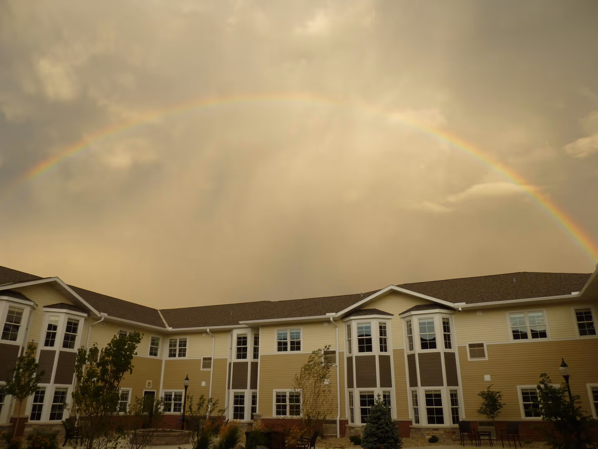 Exterior view of a two-story senior living facility building with beige and brown siding, multiple windows, and a landscaped courtyard with small trees and benches under a cloudy sky with a visible rainbow arching above.