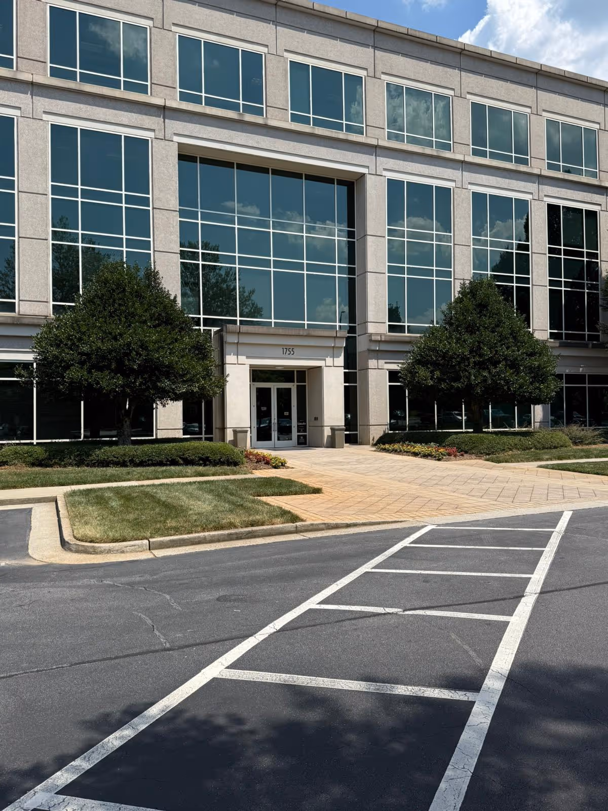Modern glass-front building exterior with entrance, landscaping, and a parking area.