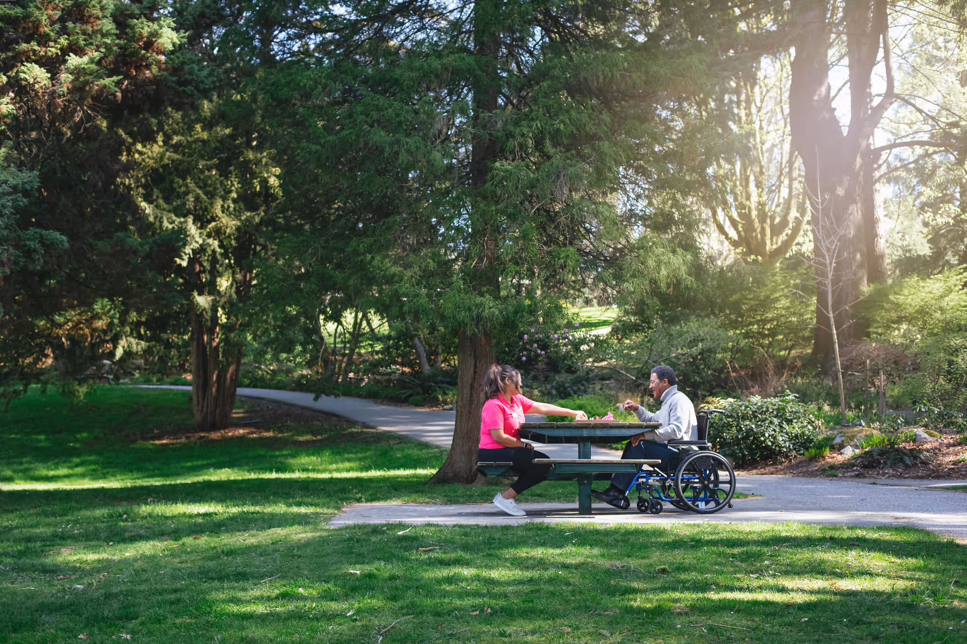 Two people, one in a wheelchair, sit at a picnic table in a sunny outdoor park.