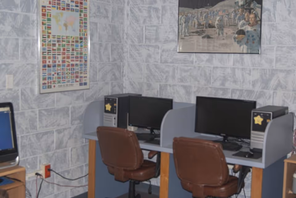 A small computer workstation area with two desktop computers on a shared desk, each with a brown swivel chair. The walls are covered with a gray stone-patterned wallpaper, and there are two framed posters hanging on the wall, one featuring world flags and the other depicting astronauts on the moon.