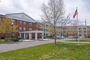 Front entrance and driveway of a red-brick senior living building with a covered porte-cochere, lawn, trees, and an American flag.