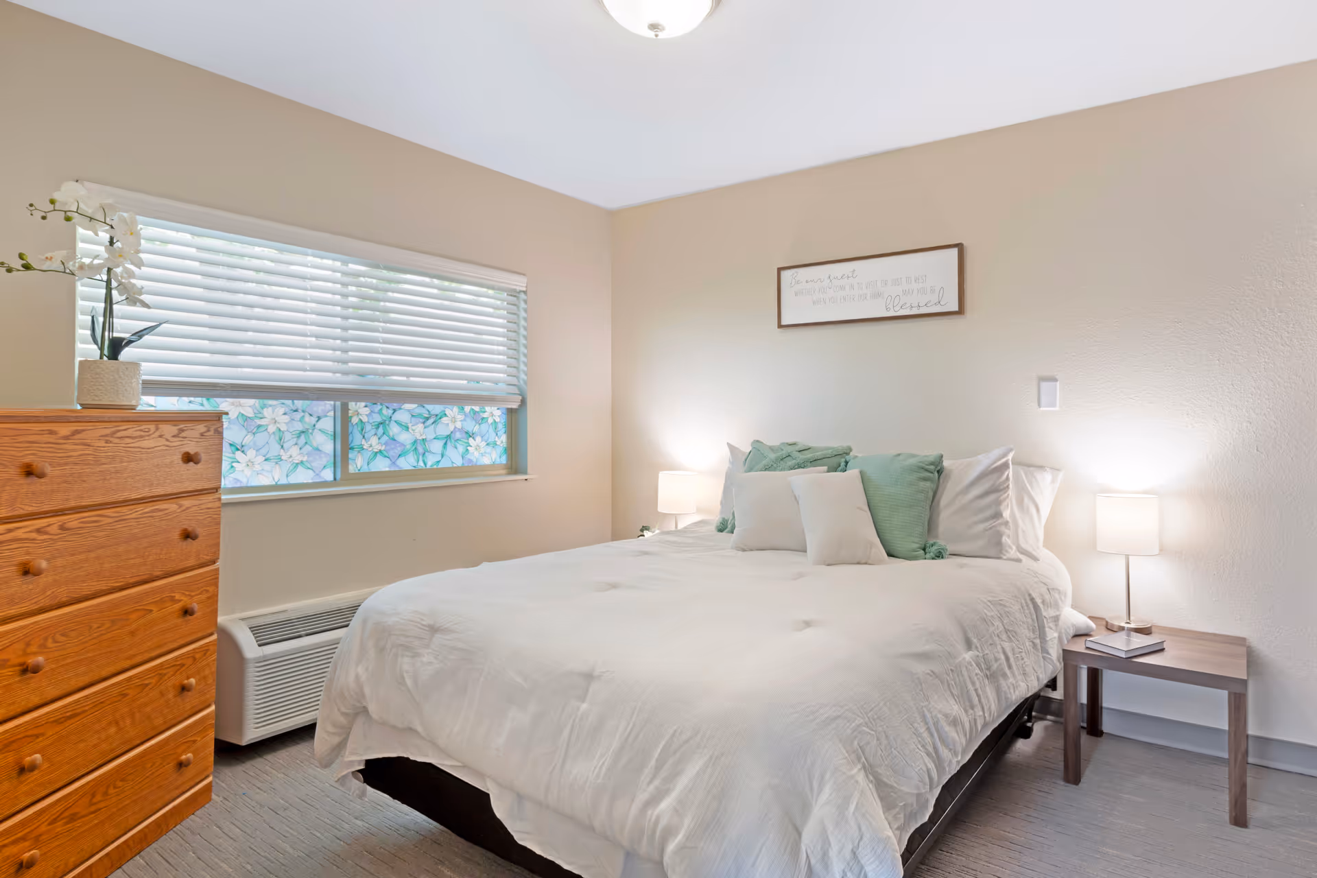 Bright bedroom with a large white bed, pillows, dresser, nightstands, and window blinds.