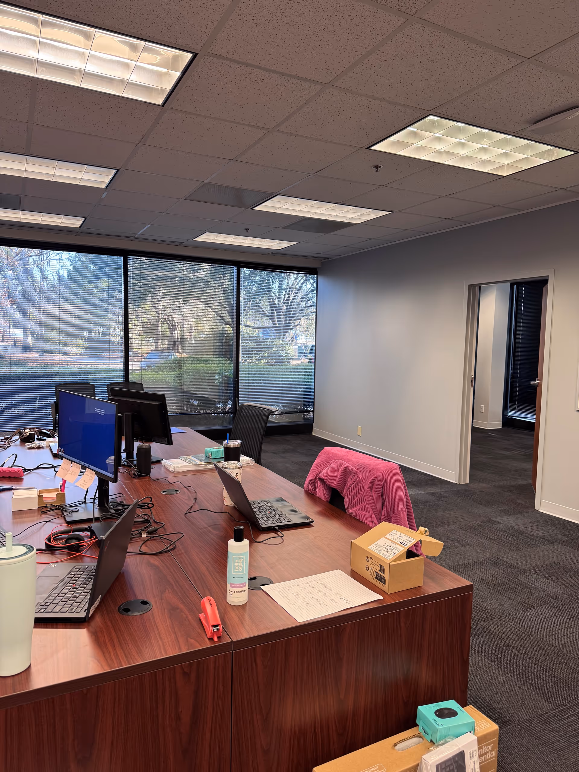 Interior office workspace with laptops, monitors, cables, and a view through large windows.