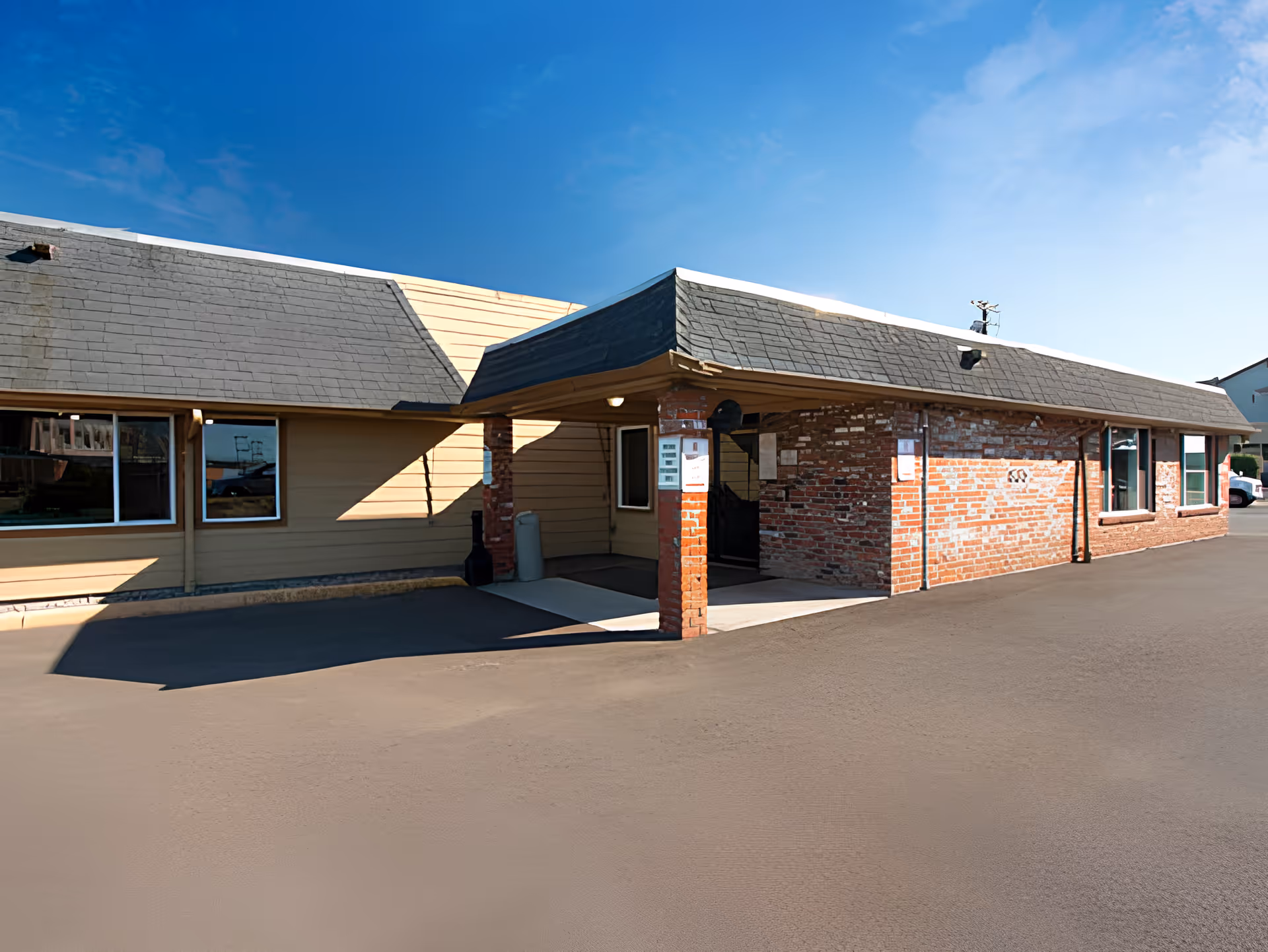 Single-story brick and siding building with a covered entrance/drive-through and asphalt parking area under a clear blue sky.