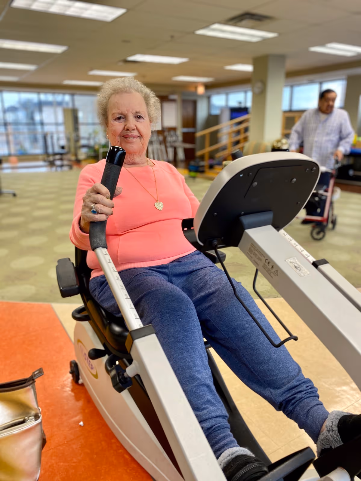 An elderly woman wearing a pink long-sleeve shirt and blue pants is exercising on a seated rowing machine in a spacious indoor area with carpeted floors and large windows. In the background, a man with a walker is visible near a staircase.