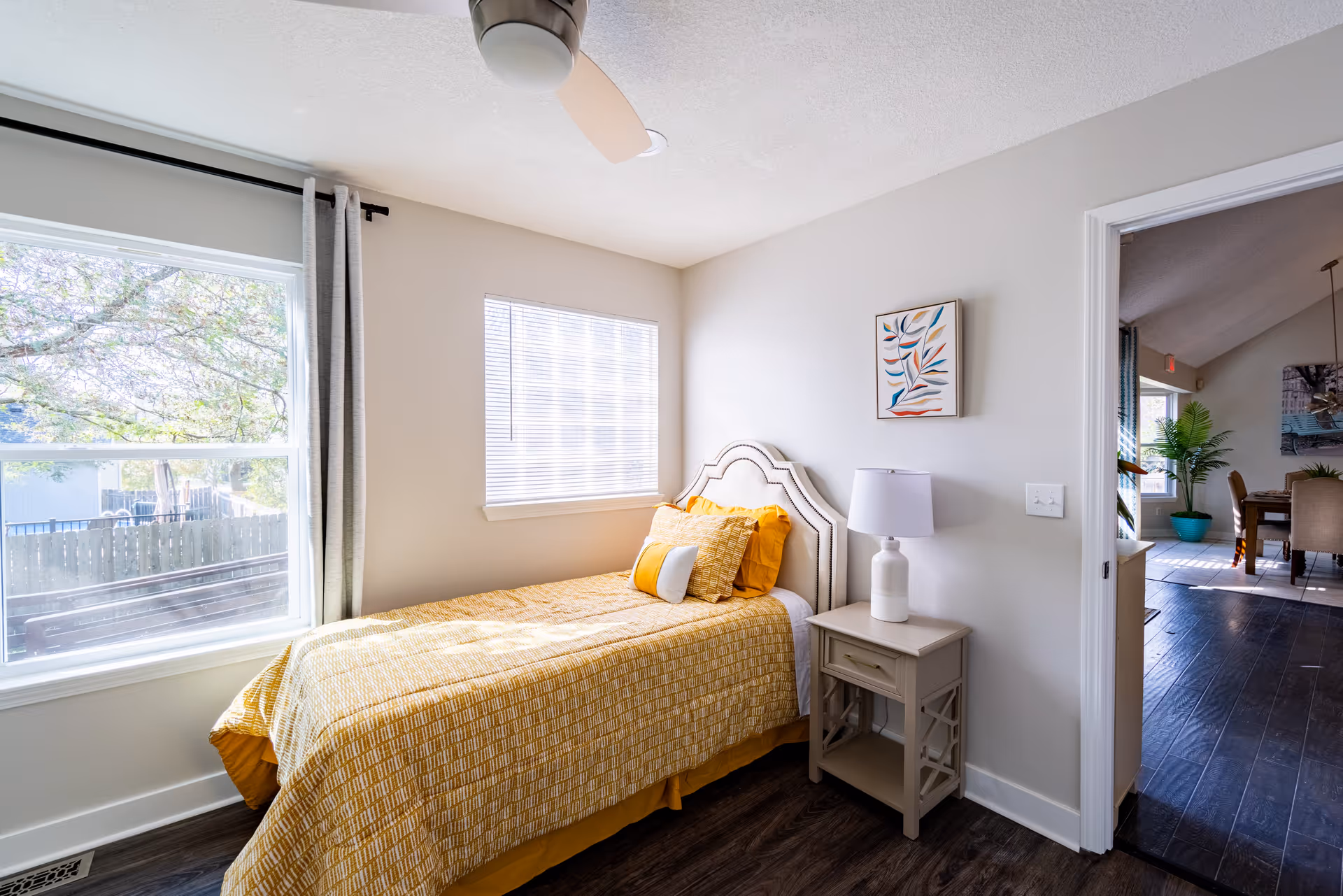 Bright bedroom with a yellow quilted bed, nightstand, lamp, and doorway to a living area.