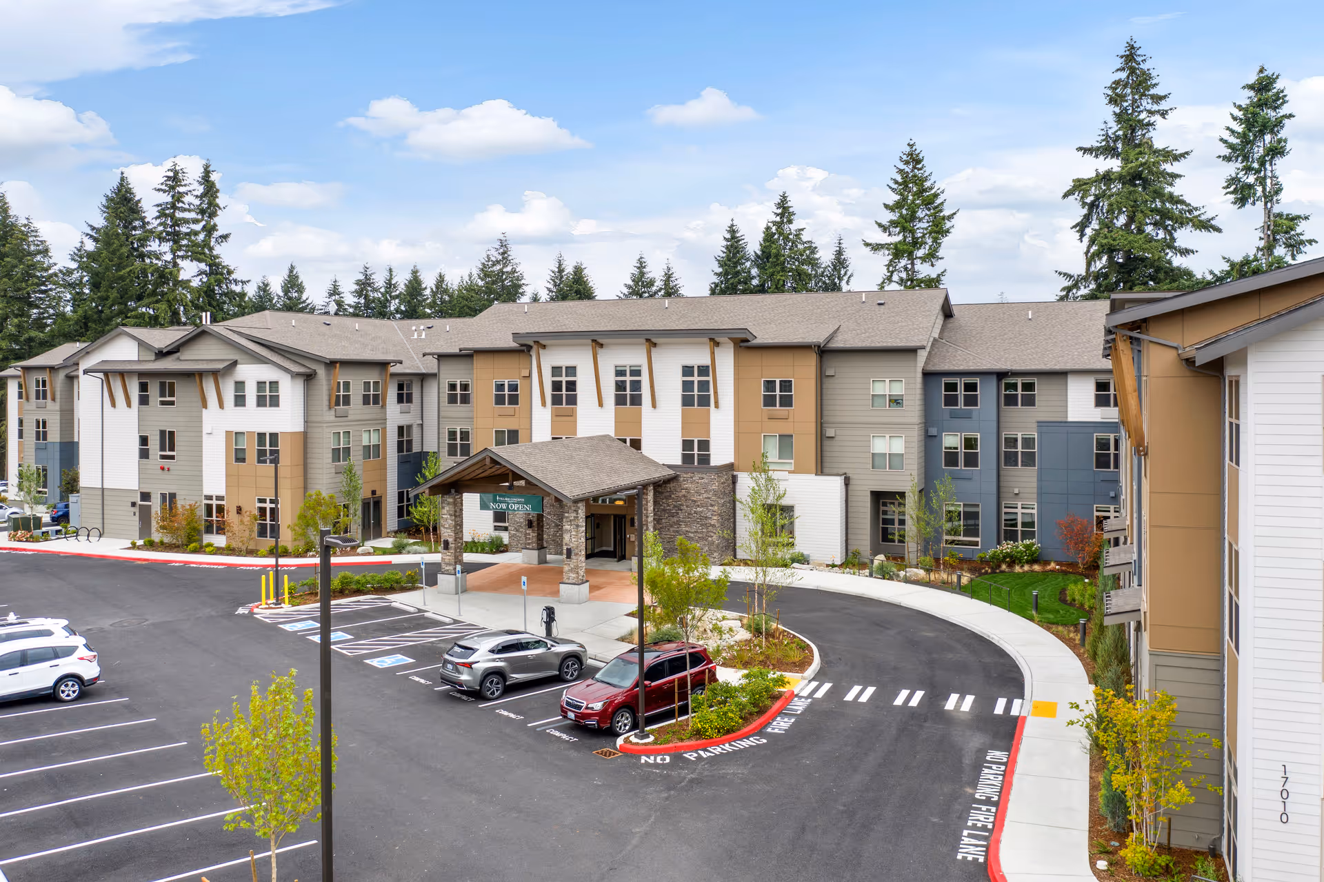 Exterior front view of a three-story senior living facility with a covered entrance, parking lot, and surrounding trees.