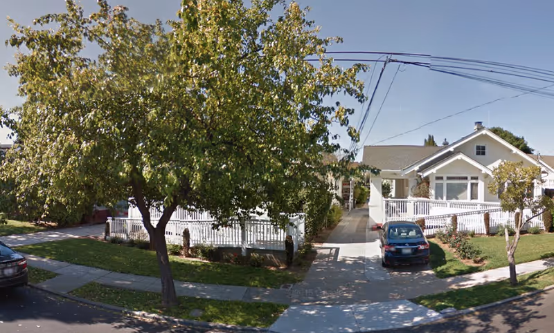 Street view of a single-story senior facility exterior with a white fence and trees.