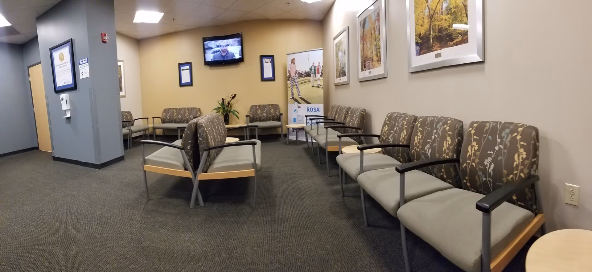 Interior waiting area with rows of cushioned chairs, framed artwork, and a wall-mounted TV.