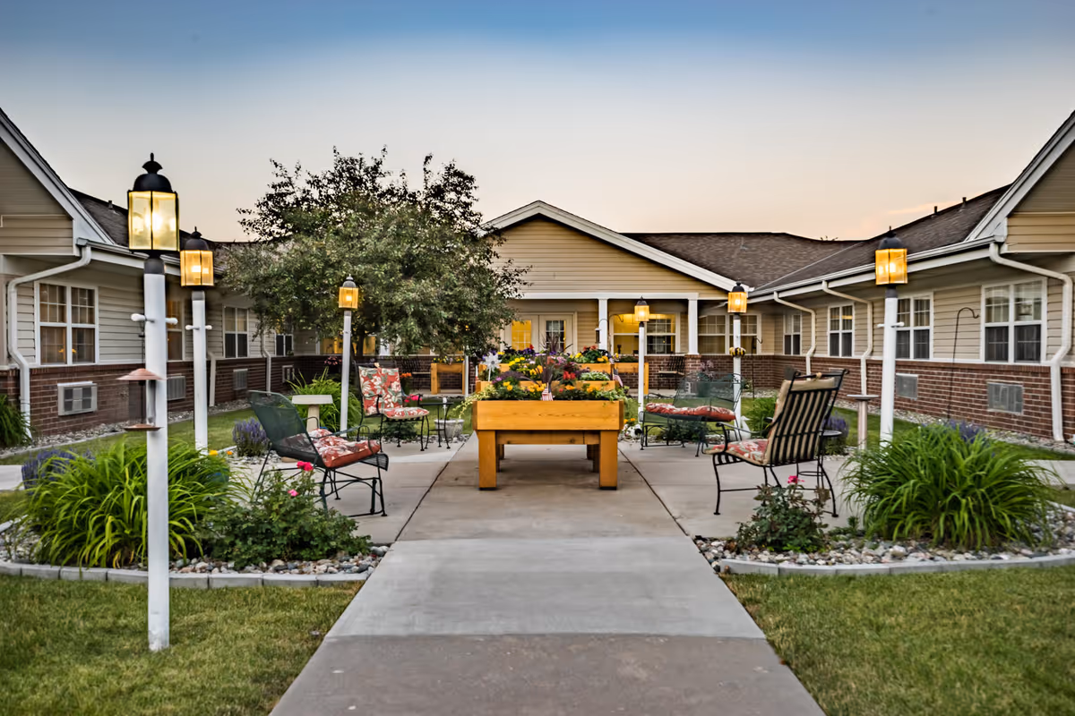 Outdoor courtyard area of a senior living facility with a paved walkway leading to a central raised garden bed filled with flowers. Surrounding the garden bed are several metal chairs with floral cushions and small tables. The courtyard is bordered by a single-story building with beige siding and brick accents. Several lamp posts with glowing lantern-style lights line the walkway and seating area. The sky is clear with a soft gradient from blue to light orange, indicating early evening.