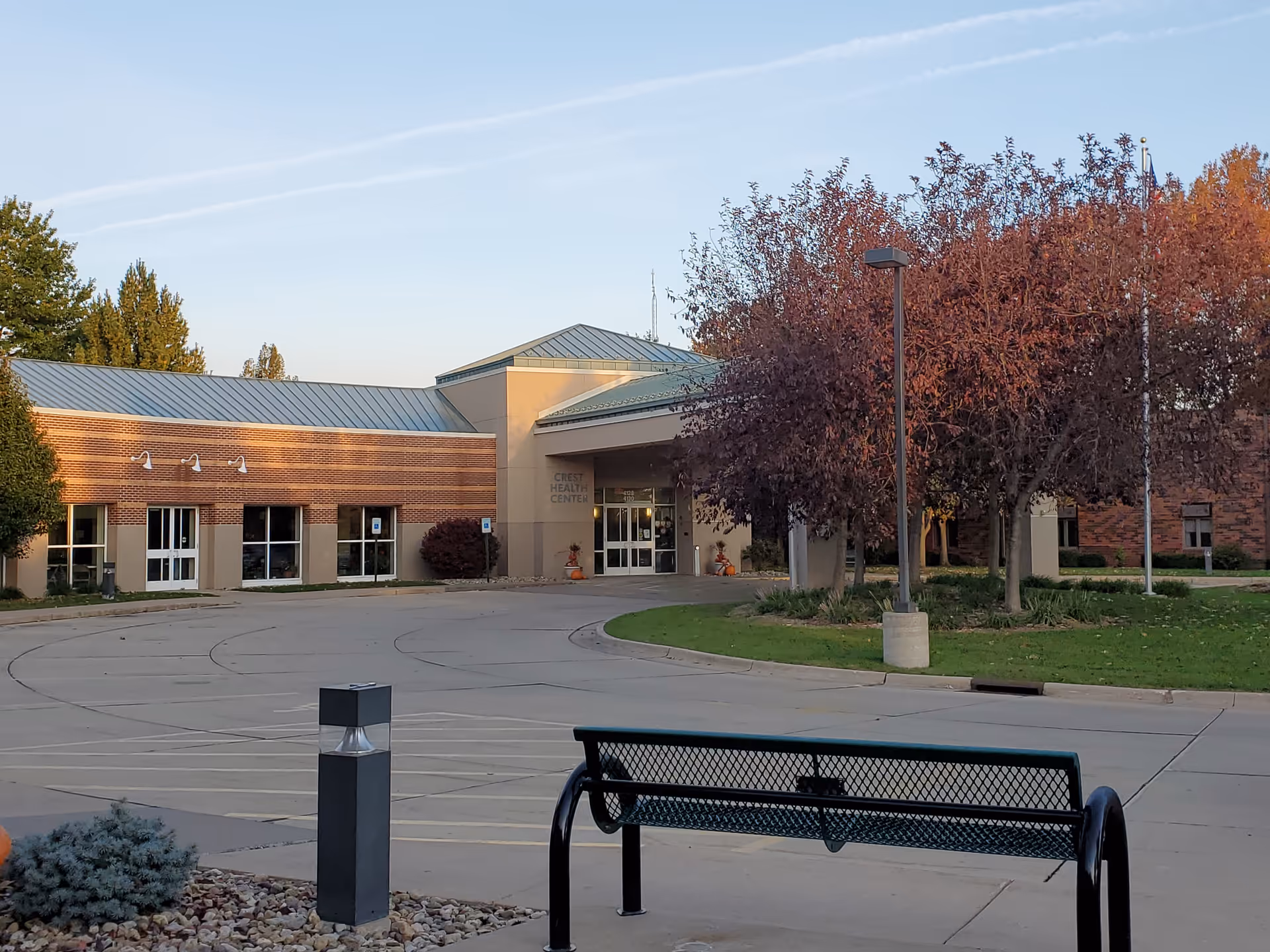 Exterior view of a single-story brick building with a metal roof and an entrance labeled Crest Health Center. There is a curved driveway in front, a black metal bench, a lamp post, and trees with autumn-colored leaves.