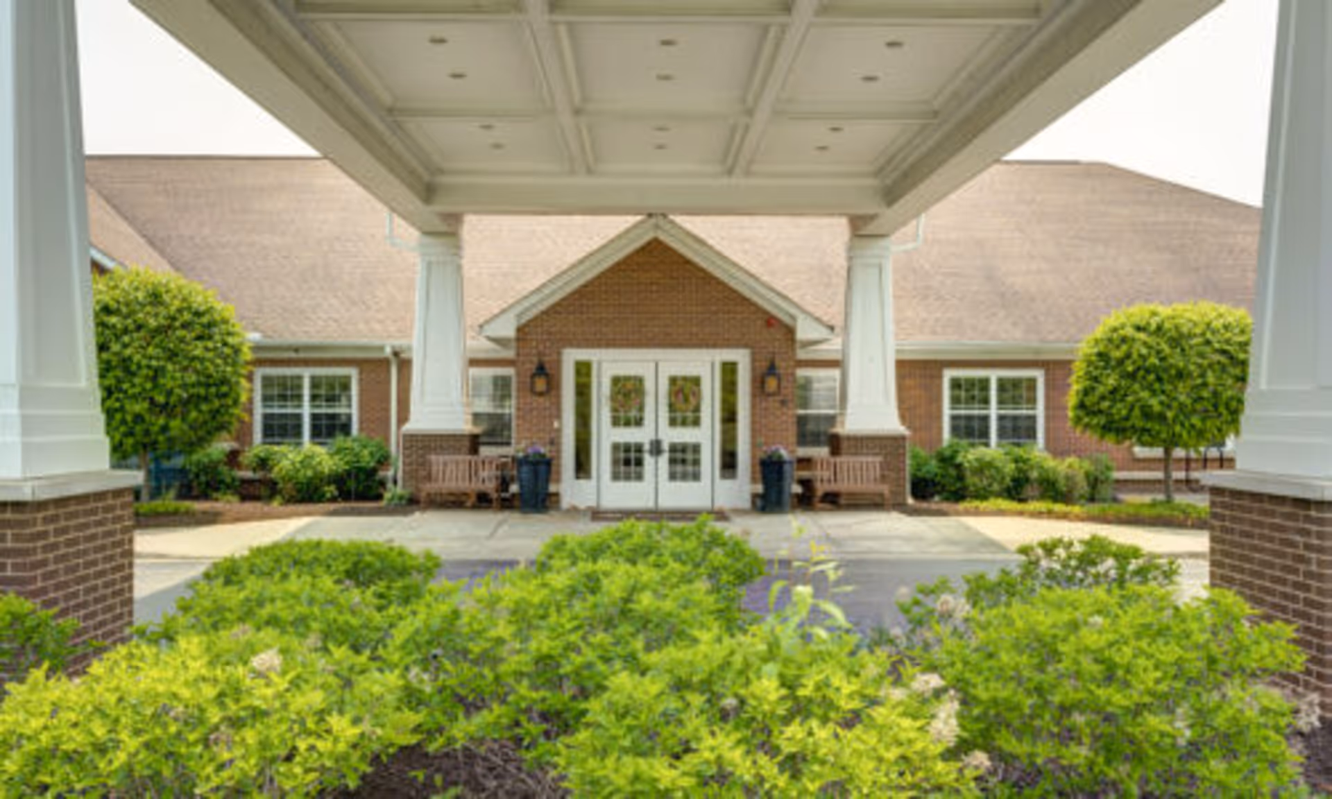 Covered porte-cochere and main entrance of a brick senior living building with double doors, benches, and landscaped shrubs in front.