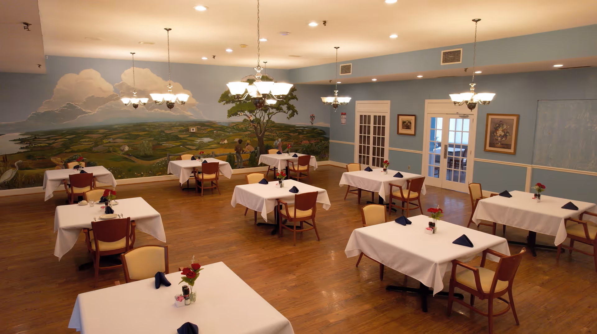Spacious dining room with white tablecloths, chandeliers, and a landscape mural.
