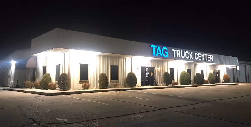 Nighttime exterior of a front-facing building with illuminated TAG Truck Center sign and trimmed shrubs.