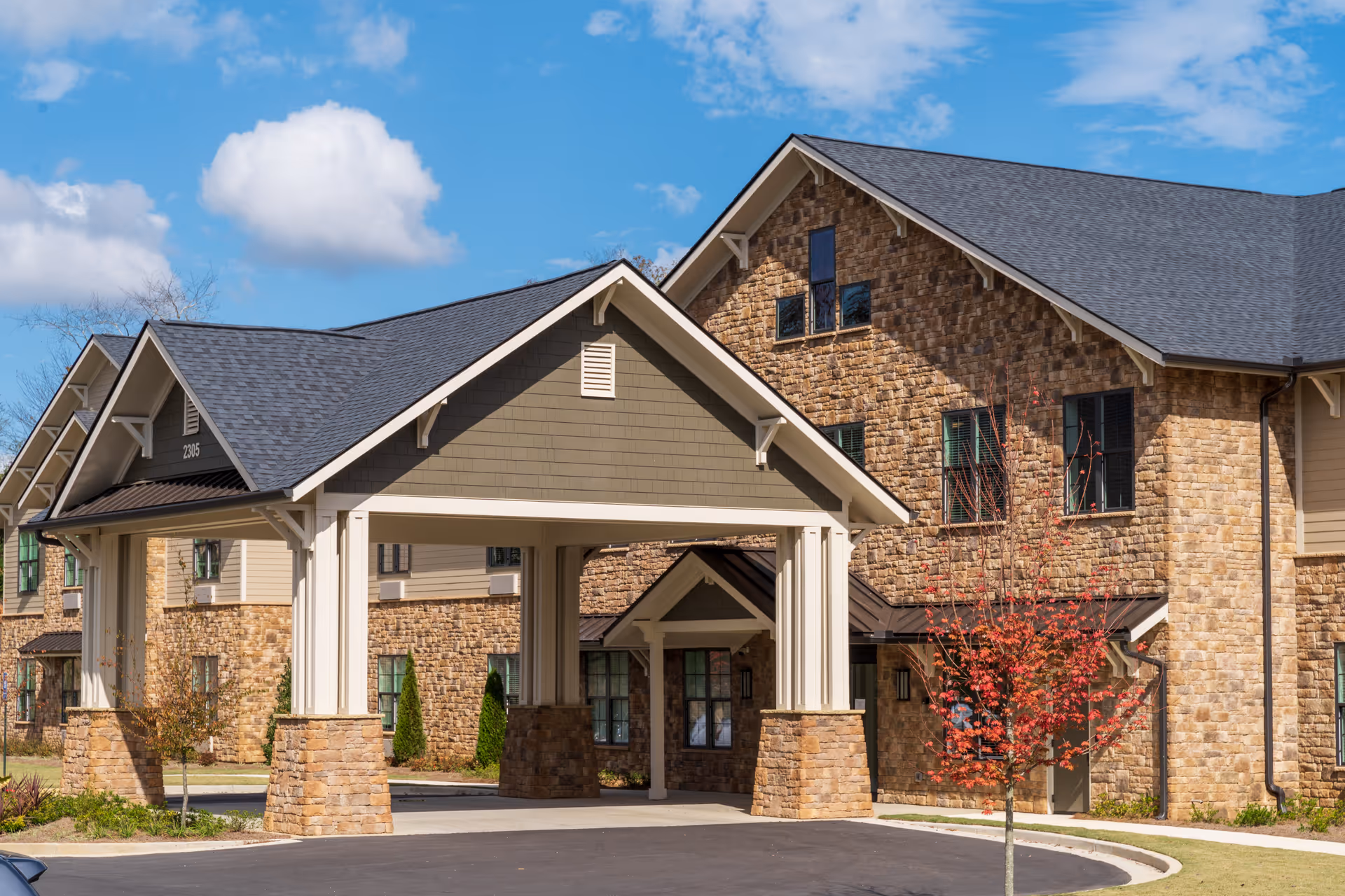 Front entrance and porte-cochere of a stone-faced senior living building with a small red-leaved tree in front.