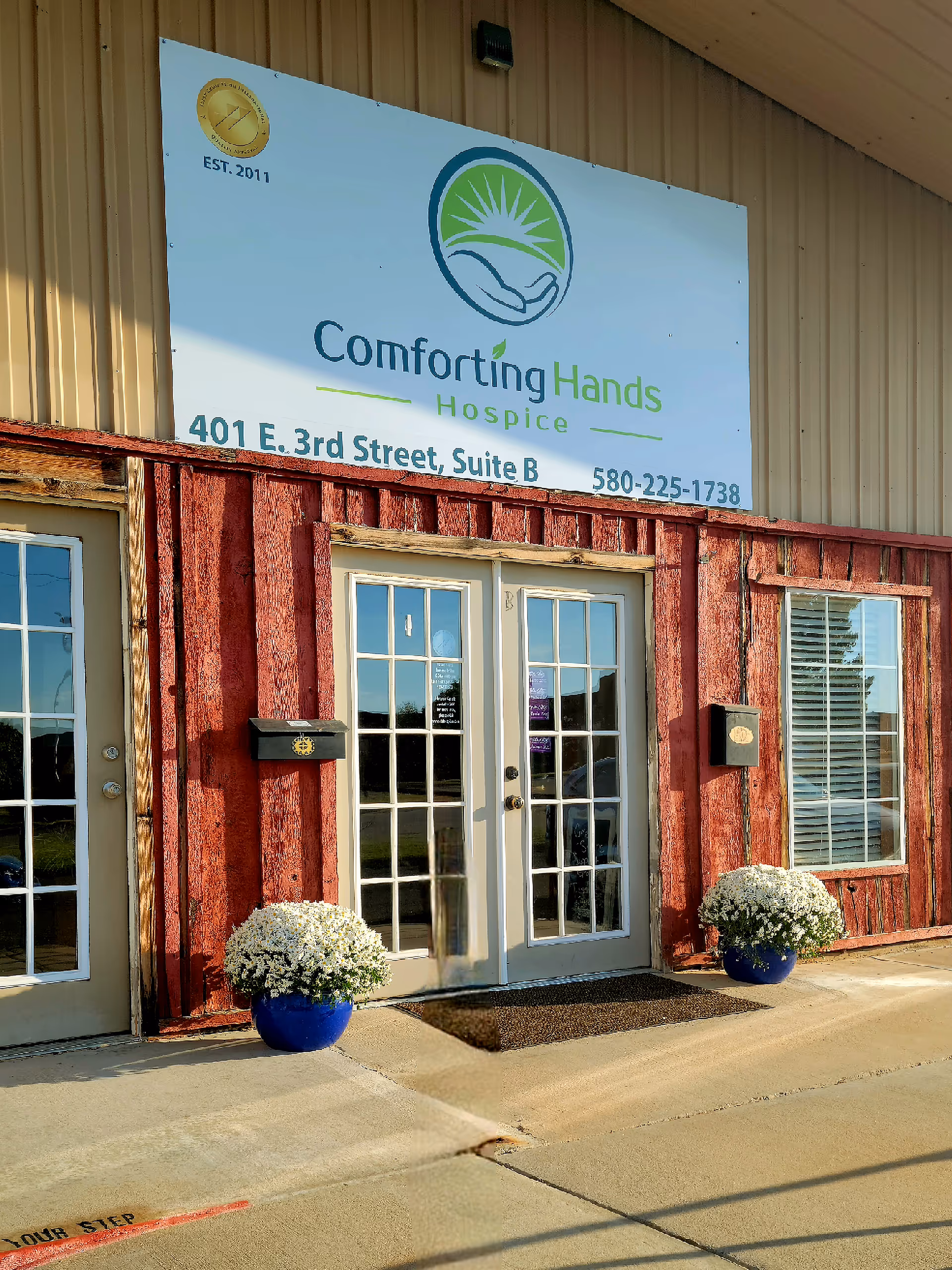 Front entrance of Comforting Hands Hospice with sign and white-potted flowers.