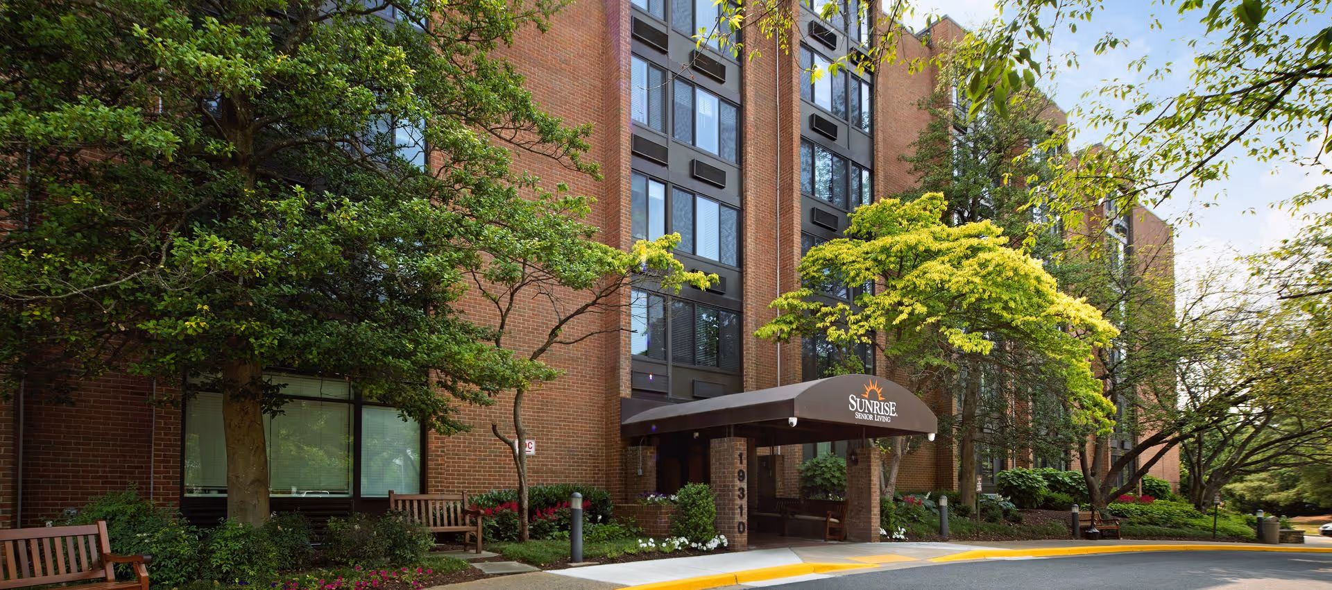Front entrance of a brick senior living building with a covered canopy reading 'Sunrise', flanked by trees, landscaping, and benches.