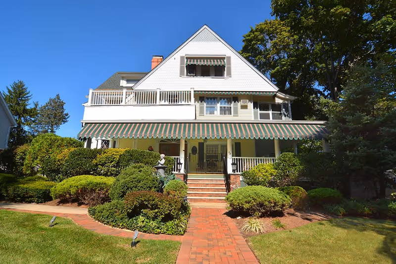 Front exterior view of a large, white, multi-story house with a green and white striped awning over the porch. The house is surrounded by well-maintained bushes and greenery, with a brick pathway leading up to the front steps. The sky is clear and blue.