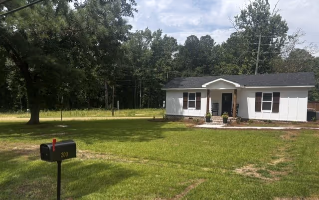 Front view of a small white house with a porch in a grassy yard with trees.