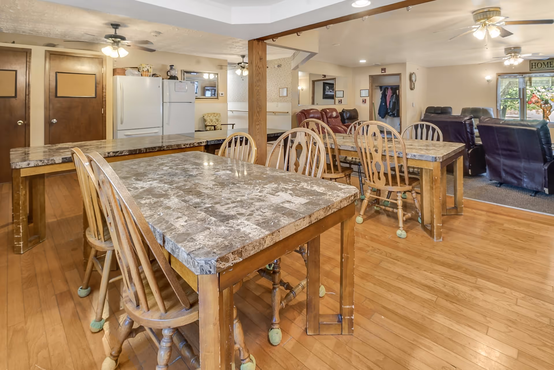 Communal dining area with multiple marble-top tables and wooden chairs, a refrigerator along the back wall and a seating/lounge area visible beyond.