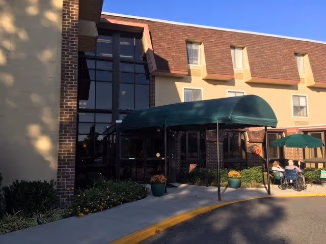 Exterior view of Ginter Hall South building entrance with a green canopy over the doorway. The building has a brick and beige facade with several windows. There are potted plants near the entrance and two people sitting outside, one in a wheelchair, under a green umbrella.